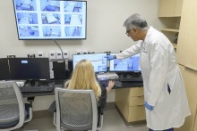 Man wearing white coat stands pointing at a screen. A woman with long blonde hair sits at a desk in front of the computer screen the man is pointing at.