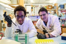 A student and professor, both wearing white lab coats, pose for a photo in a laboratory