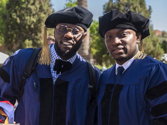 Two young men dressed in graduation regalia stand outside before convocation.