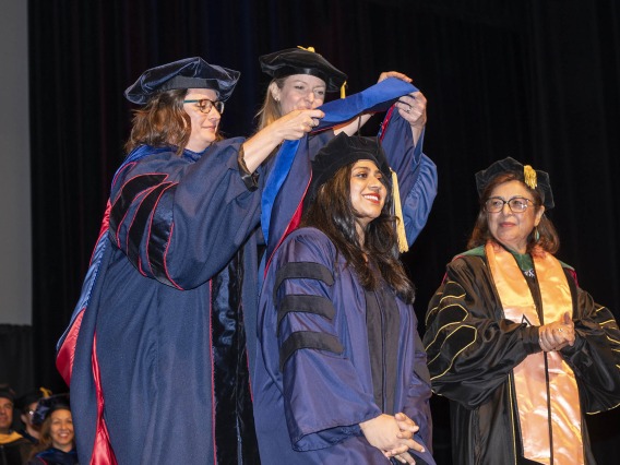Two female faculty in graduation regalia place a sash over the shouldders of a smiling female graduate while another female faculty member looks on. 