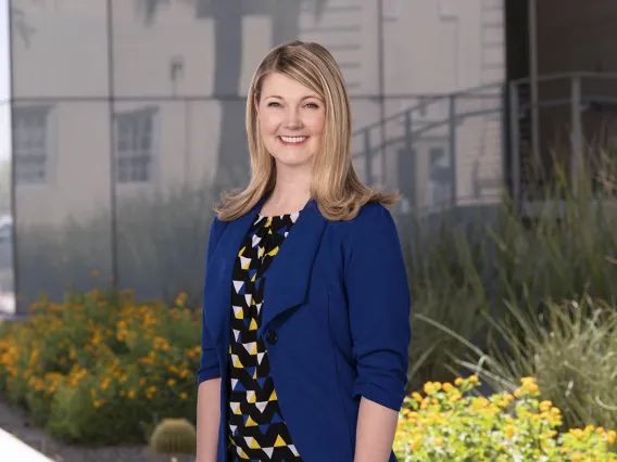 Woman with shoulder length blonde hair wearing a black, white, yellow and blue blouse with a blue blazer on top. She is smiling and standing outside.