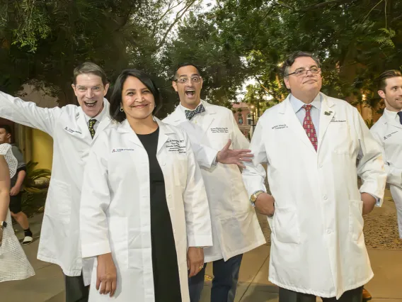 Six college of medicine faculty members wearing white coats and acting silly walk outside on their way to the white coat ceremony. 