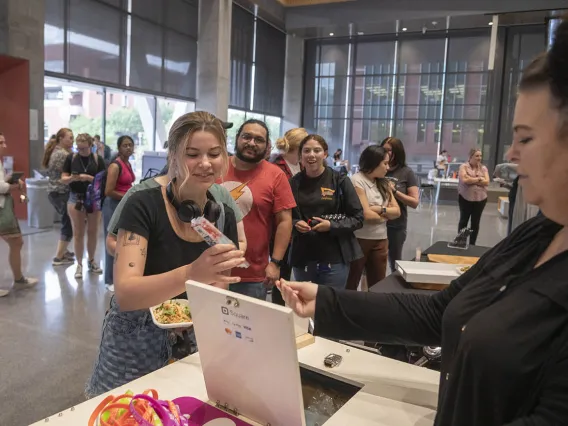 A young woman reaches for a small salad plate from a counter as dozens of other people mingle in the background. 