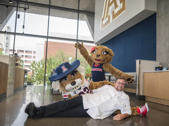 A male chef lays on his side next to the Wilbur the Wildcat mascot as the Wilma Wildcat mascot stands behind them with arms raised. 