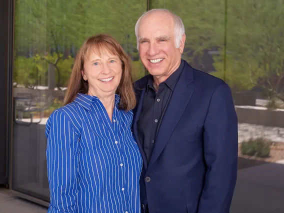 Woman with brown hair wearing a blue shirt stands with a man with grey hair wearing a black suit