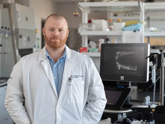 Light-skinned man with a red beard wearing a blue shirt and a white lab coat stands in a lab surrounded by equipment.