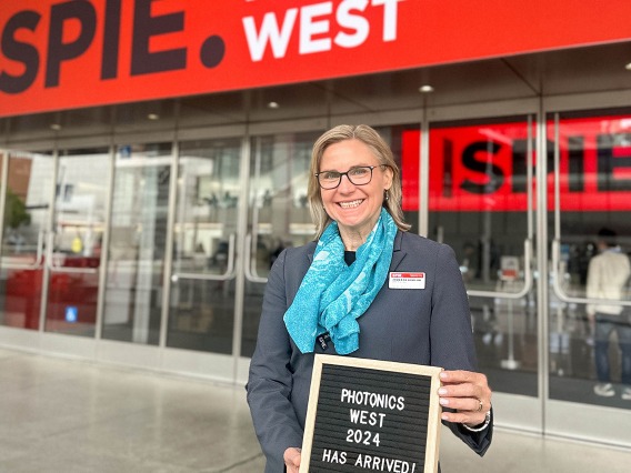 Dr. Jennifer Barton stands in front of a building that has “SPIE” on it holding a sign that reads, “Photonics West 2024 has arrived.” 