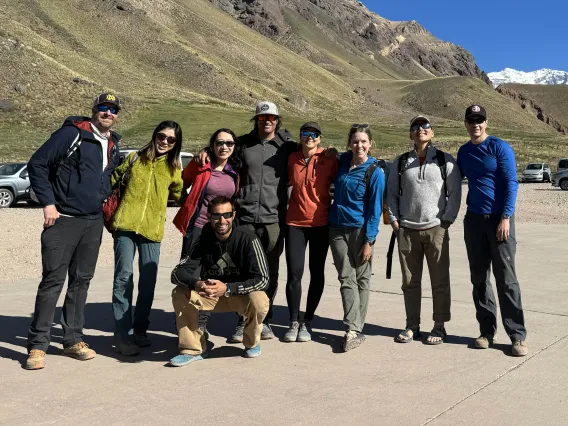 Nine people stand in a parking lot smiling with green mountains in the background. 