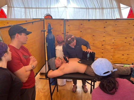 A man lies on his back on a table while Nicola Baker holds a portable ultrasound machine on his stomach in one hand and an ultrasound wand in another hand against his chest. 