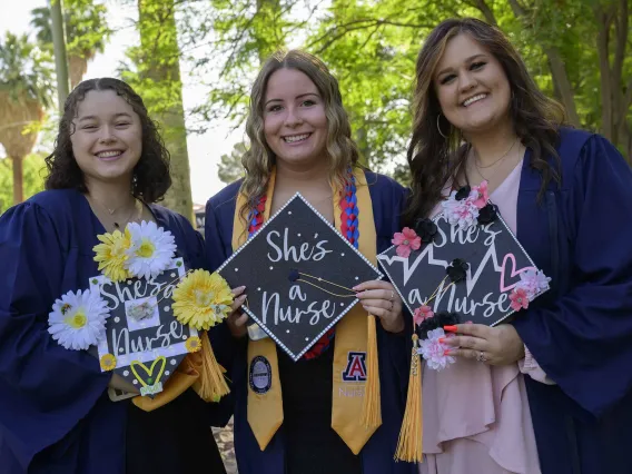 Three University of Arizona College of Nursing Bachelor of Science in Nursing students in graduation gowns show the decorated tops of their graduation caps. 