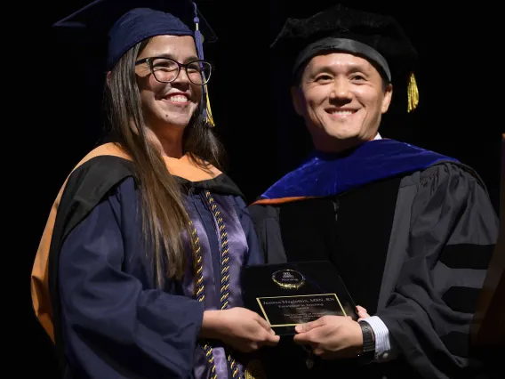 Two University of Arizona College of Nursing faculty members in graduation caps and gowns stand together smiling as they hold an award plaque in their hands. 