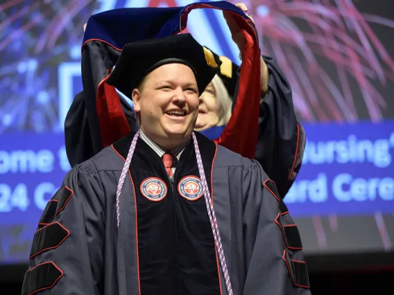 A University of Arizona College of Nursing professor places a hood over the shoulders of a nursing student. Both are dressed in graduation caps and gowns.