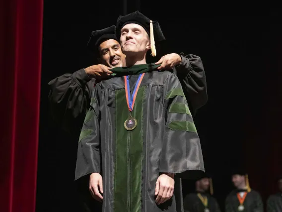 A University of Arizona College of Medicine – Tucson student dressed in a graduation cap and gown has a ceremonial hood placed over his shoulders by his faculty mentor, who is also dressed in graduation regalia.