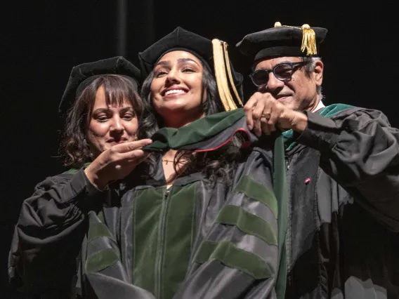 Two University of Arizona College of Medicine – Tucson professors place a ceremonial graduation hood over the shoulders of their daughter. All are dressed in caps and gowns and smiling. 