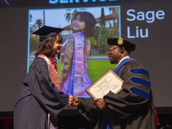 A University of Arizona Mel and Enid Zuckerman College of Public Health student shakes hands with a professor who is about to hand her an award. Both are dressed in graduation regalia. 