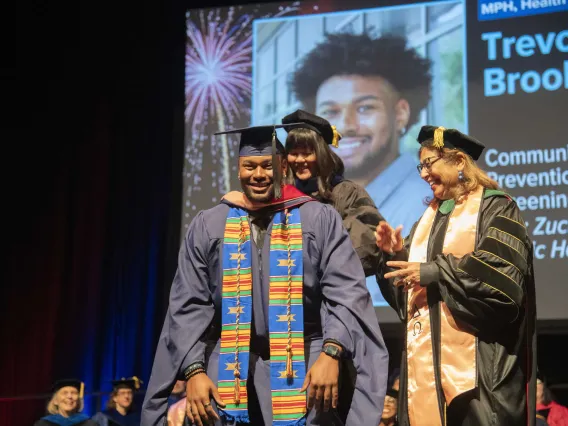 A smiling University of Arizona Mel and Enid Zuckerman College of Public Health student dressed in graduation regalia stands on a stage as two faculty members place a graduation hood over his shoulders. 