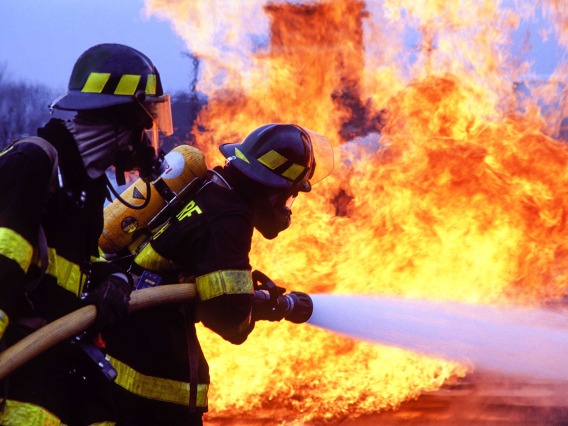Two firefighters wearing protective gear and oxygen tanks spray water onto a blaze