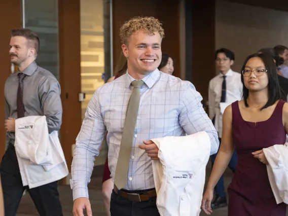 A new University of Arizona College of Medicine – Phoenix student dressed in a shirt and tie smiles as he holds a medical white coat over his arm.