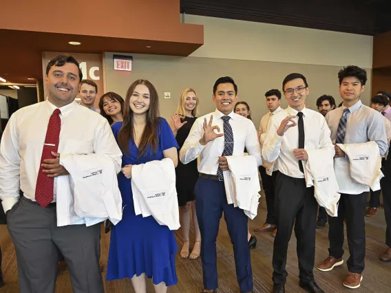 A group of new U of A College of Medicine – Phoenix medical students dressed in shirts, ties and dresses, all with medical white coats draped over their arms, stand, smile and show the hand sign for the Arizona Wildcats. 