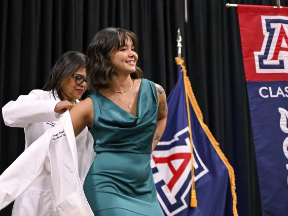 A smiling University of Arizona College of Medicine – Phoenix student in a green dress stretches out her arms as a professor puts a medical white coat on her.