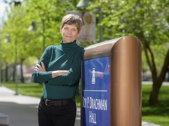 Woman with short brown hair wearing a green turtleneck and black pants leans again the University of Arizona Roy P. Drachman Hall sign