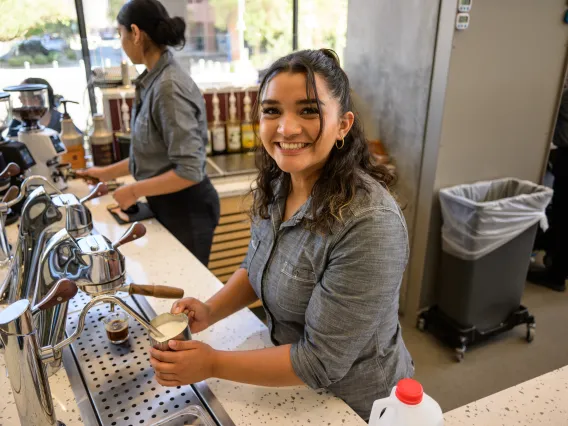 A young woman with darc curly hair froths milk at an espresso machine. 