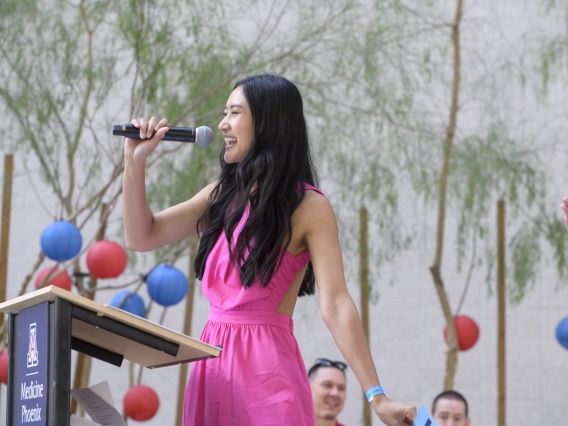A young Asian woman with long dark hair speaks into a microphone in an outdoor setting. 