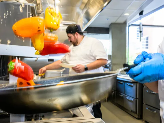 A skillet with colorful bell peppers tosses them in the air over a stove top. 