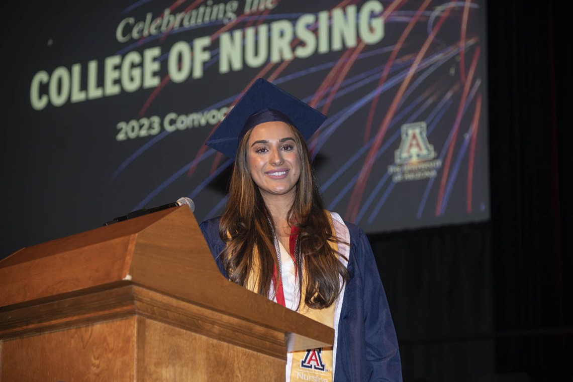 A young woman with long brown hair and dressed in cap and gown stands at a podium in Centennial Hall.