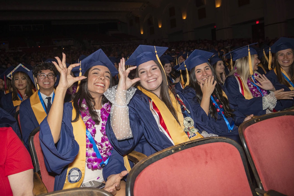 Two young women dressed in graduation regalia flash the University of Arizona hand sign while seated in Centennial Hall.