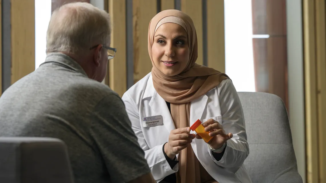 Diyana Ahmad, a second-year student at the Coit College of Pharmacy, holds a prescription bottle and discusses the medication with a male older adult patient.