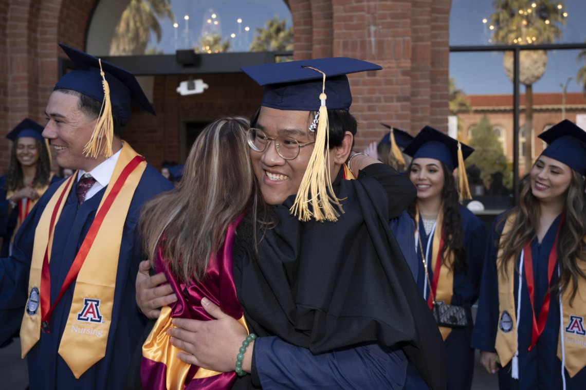 A young man in graduation cap and gown smiles as he hugs one of his professors outside. 
