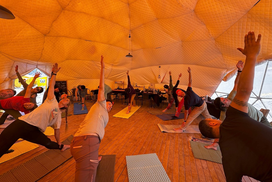 Inside a domed tent, a group of people on yoga mats lean on one arm and reach the other arm up toward the ceiling.