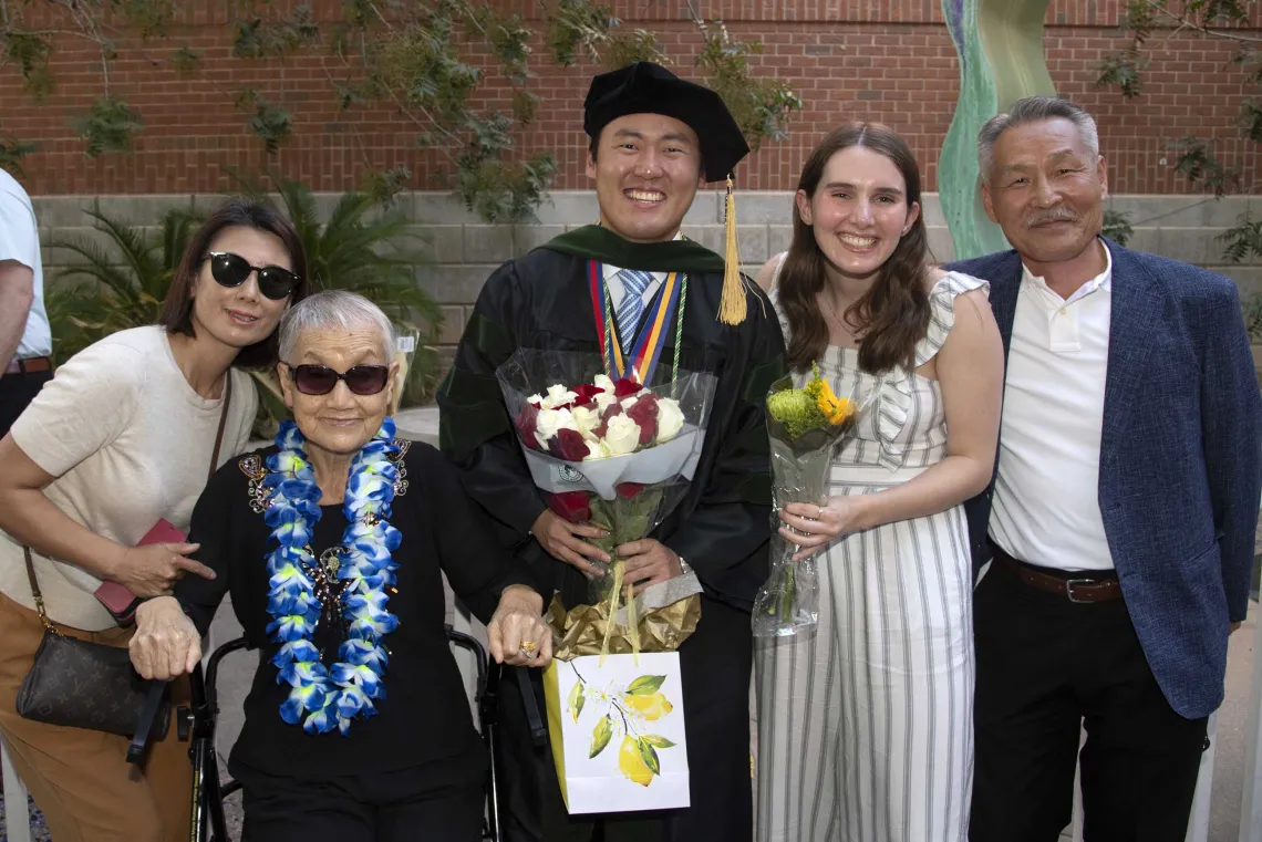 A University of Arizona College of Medicine – Tucson graduate smiles as he holds flowers and a gift bag while surrounded by family and friends. 