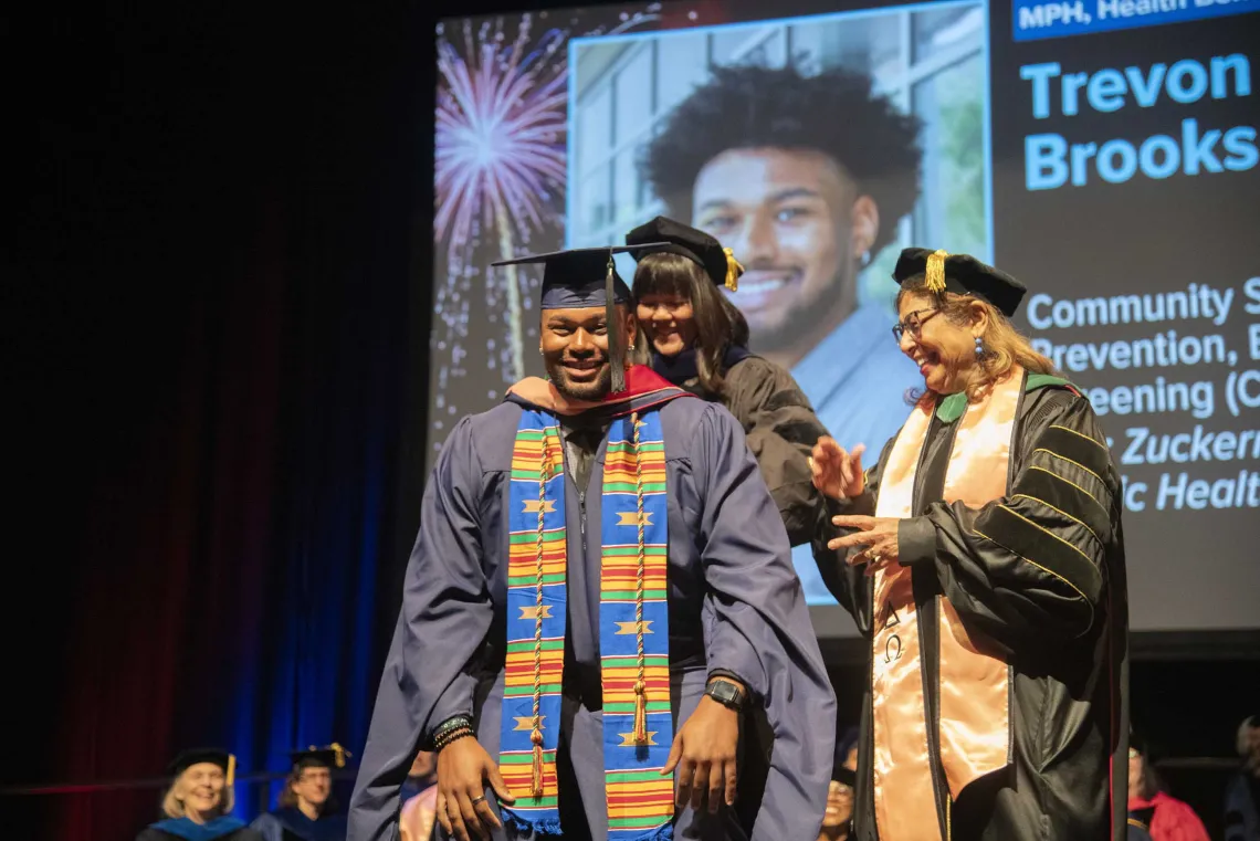 A smiling University of Arizona Mel and Enid Zuckerman College of Public Health student dressed in graduation regalia stands on a stage as two faculty members place a graduation hood over his shoulders. 