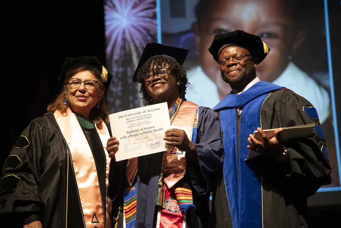 A University of Arizona Mel and Enid Zuckerman College of Public Health student holds up her diploma while standing between two faculty members. All are wearing graduation regalia. 