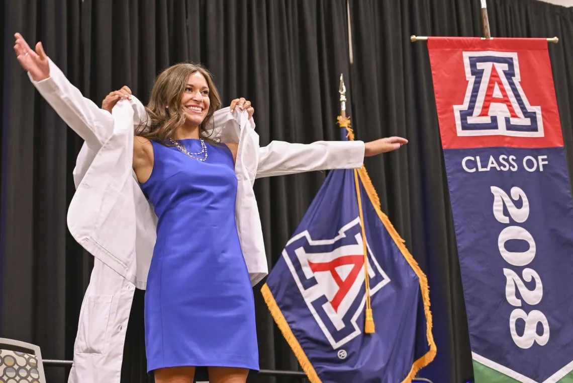 A new University of Arizona College of Medicine – Phoenix student in a blue dress stretches out her arms as a professor puts a medical white coat on her. 