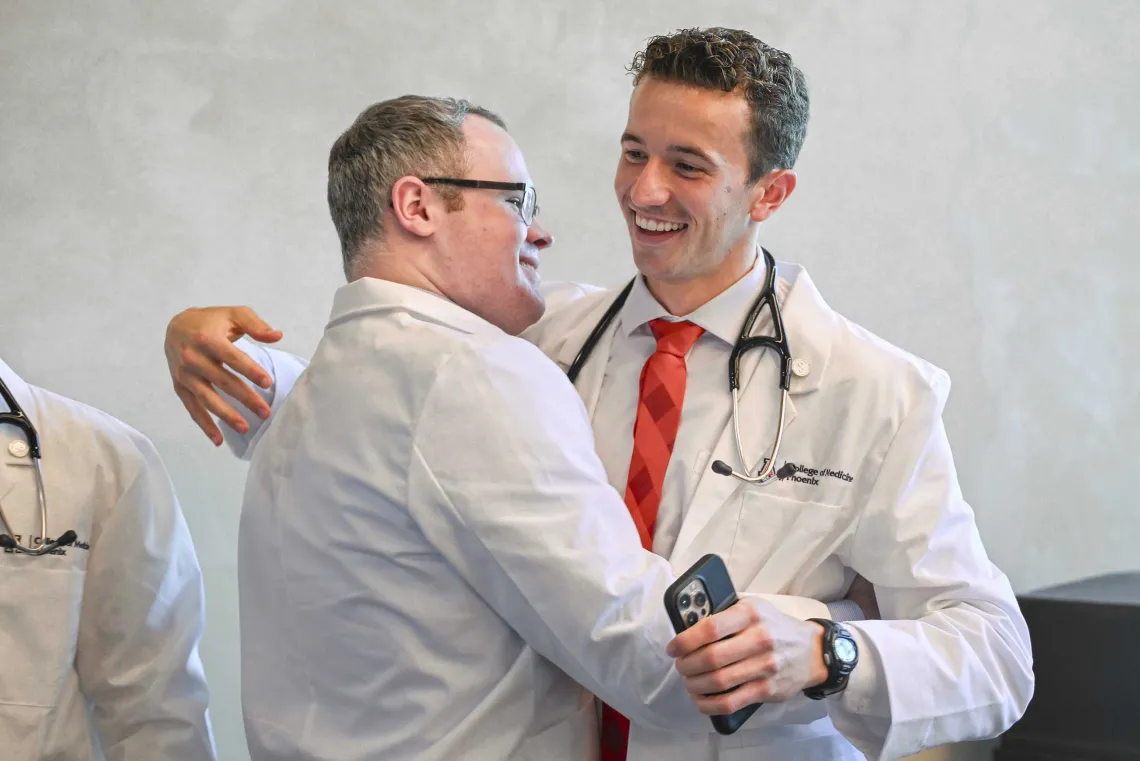 Two University of Arizona College of Medicine – Phoenix students wearing medical white coats smile as they hug. 