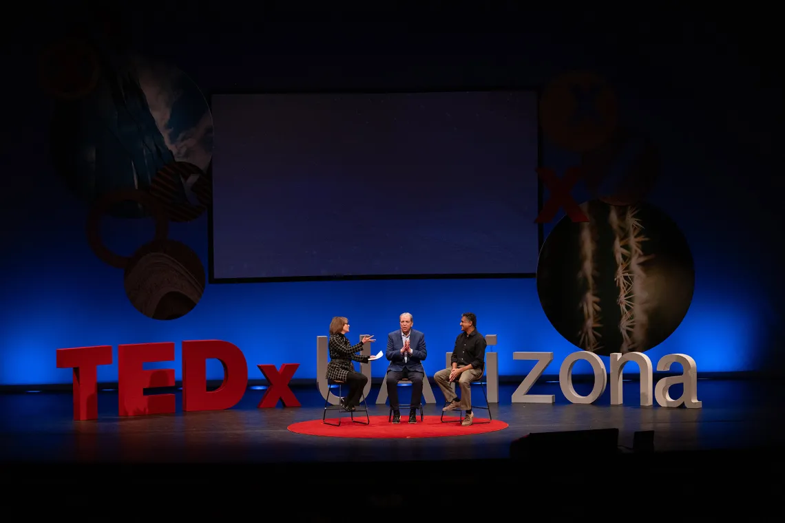 Three people sit on a stage in front of a large sign that says “UArizona.”