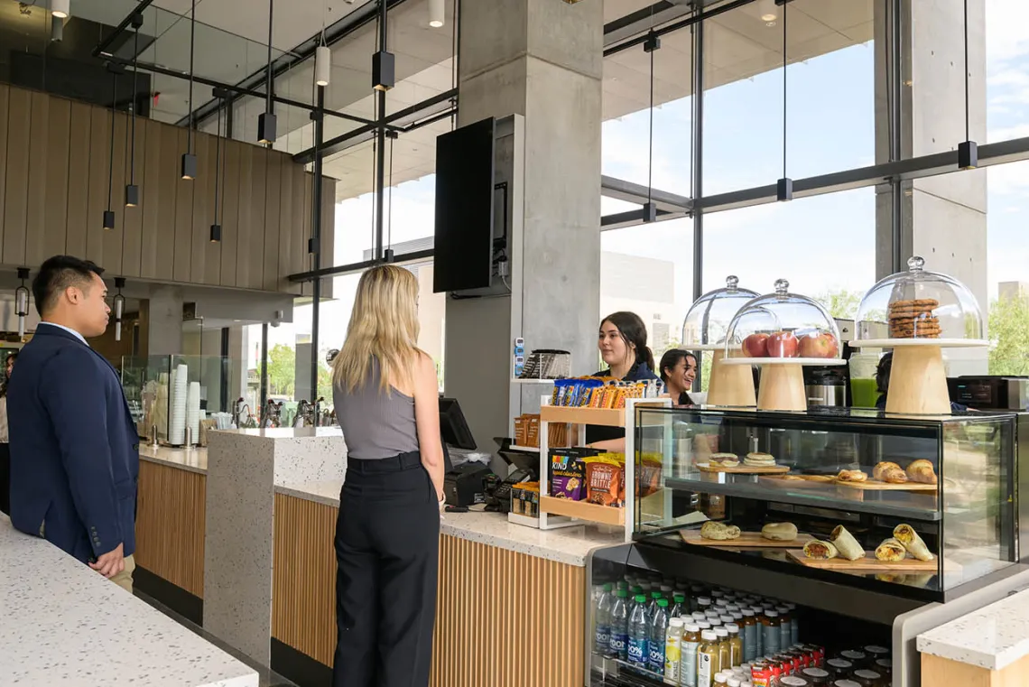 A woman with long blonde hair stands at a coffee bar counter with pastries in a case. 