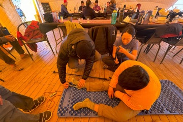 A man sits on the floor with his leg straight out while a person is bent over him wrapping something around his foot and a woman sits next to him.