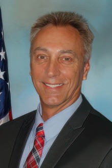 Middle-aged man with dark grey hair wearing a black suit jacket, blue shirt and red tie stands in front of a United States of America flag.